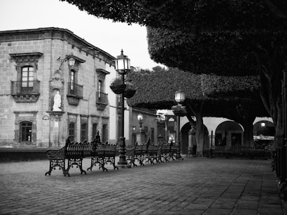 A picturesque plaza features cobblestone pavement and a series of decorative benches lining the walkway. Large, carefully trimmed trees provide shade, and vintage-style lampposts are spaced along the path. A historical building with ornate windows and balconies stands prominently in the background.