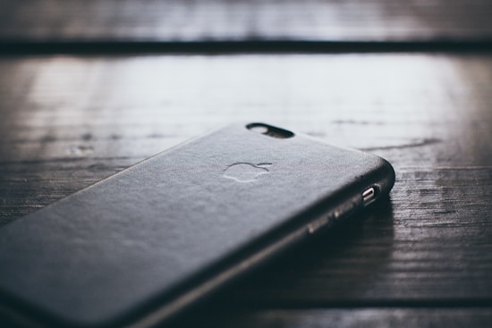 A close-up of a smartphone with a dark, textured case lying on a wooden surface. The phone's logo is faintly visible on the case.