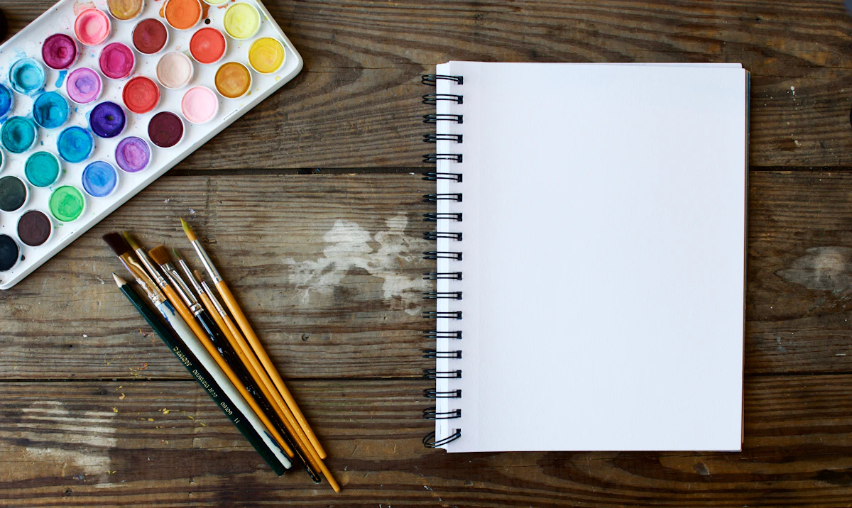 A watercolour palette and paintbrushes beside a blank sketchbook.