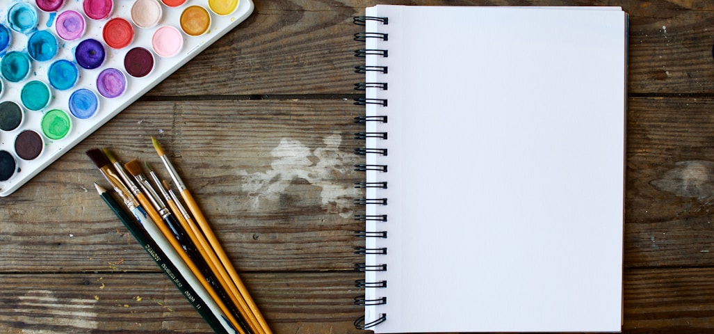 paint brushes next to drawing book and water color palette