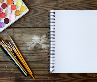 paint brushes next to drawing book and water color palette