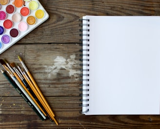 paint brushes next to drawing book and water color palette