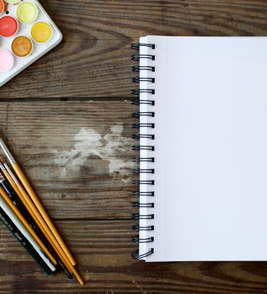 paint brushes next to drawing book and water color palette