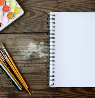 paint brushes next to drawing book and water color palette