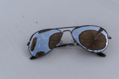 A pair of aviator sunglasses lies on a textured white surface. The reflective lenses capture the image of wooden thatched beach umbrellas set against a partly cloudy sky.