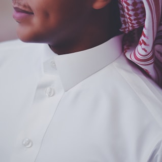 A close-up view of a person wearing a white traditional thobe with a neatly buttoned collar. The person is also wearing a patterned red and white headscarf called a keffiyeh or ghutra, draped around the neck. The image focuses on the upper torso and part of the face, showing a subtle smile and a dimple.