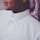 A close-up view of a person wearing a white traditional thobe with a neatly buttoned collar. The person is also wearing a patterned red and white headscarf called a keffiyeh or ghutra, draped around the neck. The image focuses on the upper torso and part of the face, showing a subtle smile and a dimple.