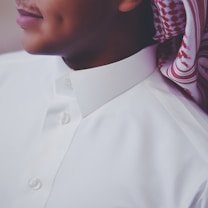 A close-up view of a person wearing a white traditional thobe with a neatly buttoned collar. The person is also wearing a patterned red and white headscarf called a keffiyeh or ghutra, draped around the neck. The image focuses on the upper torso and part of the face, showing a subtle smile and a dimple.