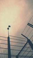 Tall, imposing metal fences angle upwards against a cloudy sky, creating a sense of enclosement and security. A security camera is mounted on a pole, overseeing the area.