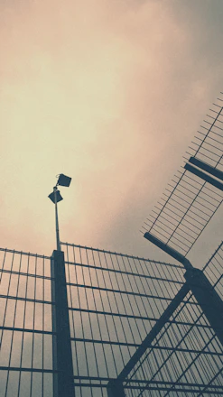 Tall, imposing metal fences angle upwards against a cloudy sky, creating a sense of enclosement and security. A security camera is mounted on a pole, overseeing the area.