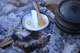 A metal pan containing a knife, a wooden cooking spoon, and a piece of yellow fruit sits among ashes and charred wood in a rustic outdoor setting. A cast iron pot is nearby, surrounded by stones and grey ashes, likely from a previous fire, suggesting a campsite cooking area.