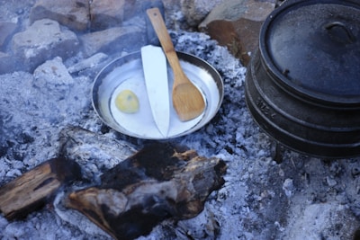 A metal pan containing a knife, a wooden cooking spoon, and a piece of yellow fruit sits among ashes and charred wood in a rustic outdoor setting. A cast iron pot is nearby, surrounded by stones and grey ashes, likely from a previous fire, suggesting a campsite cooking area.