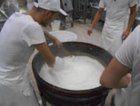 Several individuals in white uniforms are gathered around a large vat, stirring a creamy substance that appears to be cheese or yogurt. They are likely involved in a food production process. The environment is a factory or dairy setting with tiled flooring and industrial equipment in the background.