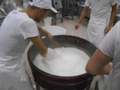 Several individuals in white uniforms are gathered around a large vat, stirring a creamy substance that appears to be cheese or yogurt. They are likely involved in a food production process. The environment is a factory or dairy setting with tiled flooring and industrial equipment in the background.
