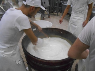 Several individuals in white uniforms are gathered around a large vat, stirring a creamy substance that appears to be cheese or yogurt. They are likely involved in a food production process. The environment is a factory or dairy setting with tiled flooring and industrial equipment in the background.