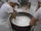 Several individuals in white uniforms are gathered around a large vat, stirring a creamy substance that appears to be cheese or yogurt. They are likely involved in a food production process. The environment is a factory or dairy setting with tiled flooring and industrial equipment in the background.