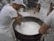 Several individuals in white uniforms are gathered around a large vat, stirring a creamy substance that appears to be cheese or yogurt. They are likely involved in a food production process. The environment is a factory or dairy setting with tiled flooring and industrial equipment in the background.
