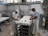 Two workers are in a cheese factory, handling trays filled with white containers. The room features industrial equipment and tiled walls, giving a clean and organized appearance. Both individuals are wearing white aprons and caps as they work.