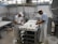 Two workers are in a cheese factory, handling trays filled with white containers. The room features industrial equipment and tiled walls, giving a clean and organized appearance. Both individuals are wearing white aprons and caps as they work.