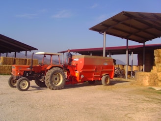 A red tractor is parked in an agricultural setting, connected to a large orange farming trailer. The background features stacks of hay bales under a metal-roofed structure, with a person standing on the trailer.