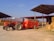 A red tractor is parked in an agricultural setting, connected to a large orange farming trailer. The background features stacks of hay bales under a metal-roofed structure, with a person standing on the trailer.