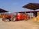 A red tractor is parked in an agricultural setting, connected to a large orange farming trailer. The background features stacks of hay bales under a metal-roofed structure, with a person standing on the trailer.