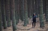 A person stands amidst a dense forest of tall, closely packed pine trees. The forest floor is covered with a layer of dry, brown pine needles. The person is wearing a dark outfit and a headband made of leaves, appearing contemplative with a peaceful expression.