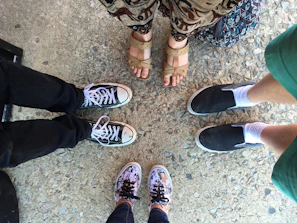 Four pairs of feet positioned in a circle, standing on textured concrete. Each person wears different footwear including black and white sneakers, beige sandals, black slip-ons with white socks, and colorful floral sneakers. The surrounding attire includes denim, a patterned skirt, and green shorts.