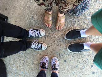 Four pairs of feet positioned in a circle, standing on textured concrete. Each person wears different footwear including black and white sneakers, beige sandals, black slip-ons with white socks, and colorful floral sneakers. The surrounding attire includes denim, a patterned skirt, and green shorts.