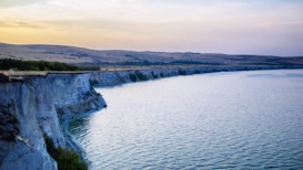 A serene landscape featuring a long stretch of cliffs bordering a calm body of water during sunset. The cliffs are rugged and partially covered with vegetation, while the water reflects the soft light of dusk. The sky displays a gradient of pale blues and yellows, adding to the tranquil atmosphere.