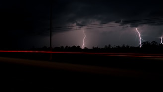 Lightning bolts illuminate the dark sky over a silhouetted forest, with red streaks of light from passing vehicles and power lines visible.