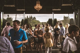 A diverse group of people is gathered in an indoor space with large windows, engaging in conversation and enjoying refreshments. The setting appears to be a social or networking event, with individuals wearing casual to semi-formal attire. A chandelier hangs from the ceiling, and natural light filters through the windows, revealing greenery outside.
