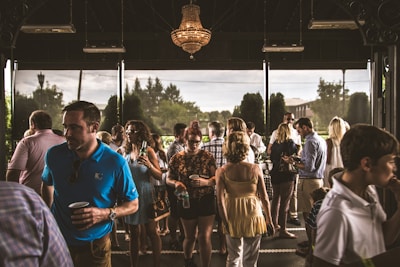 A diverse group of people is gathered in an indoor space with large windows, engaging in conversation and enjoying refreshments. The setting appears to be a social or networking event, with individuals wearing casual to semi-formal attire. A chandelier hangs from the ceiling, and natural light filters through the windows, revealing greenery outside.