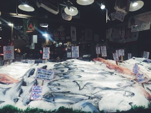 A seafood market display with various types of fish laid out on a bed of ice. Multiple handwritten signs indicate the types of fish and their prices, including wild salmon and mackerel. The background shows hanging lights and various signs, contributing to the bustling market atmosphere.
