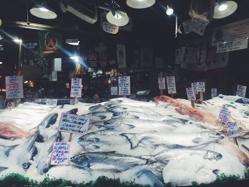 A seafood market display with various types of fish laid out on a bed of ice. Multiple handwritten signs indicate the types of fish and their prices, including wild salmon and mackerel. The background shows hanging lights and various signs, contributing to the bustling market atmosphere.