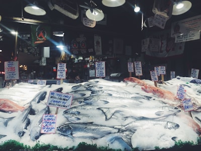 A seafood market display with various types of fish laid out on a bed of ice. Multiple handwritten signs indicate the types of fish and their prices, including wild salmon and mackerel. The background shows hanging lights and various signs, contributing to the bustling market atmosphere.