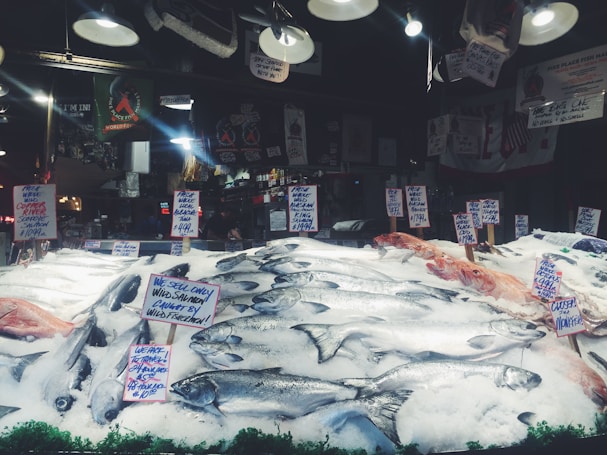 A seafood market display with various types of fish laid out on a bed of ice. Multiple handwritten signs indicate the types of fish and their prices, including wild salmon and mackerel. The background shows hanging lights and various signs, contributing to the bustling market atmosphere.