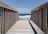 A wooden boardwalk leads out towards the ocean, with a clear view of the waves gently crashing against the shore. The sky is bright and blue, complementing the deep blue of the water. Wooden railings line the walkway, casting shadows on the planks.