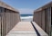 A wooden boardwalk leads out towards the ocean, with a clear view of the waves gently crashing against the shore. The sky is bright and blue, complementing the deep blue of the water. Wooden railings line the walkway, casting shadows on the planks.