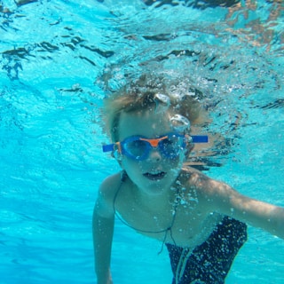 A young child, partially submerged underwater, swimming with blue and orange goggles. Bubbles can be seen around the child’s face, and the water is clear, showcasing different shades of blue.