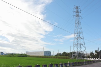 A large metal transmission tower stands tall against a partly cloudy sky, surrounded by green fields and a distant industrial building. Power lines stretch across the sky, and a row of short barriers lines the edge of a road in the foreground.