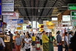 A bustling indoor market with numerous jewelry shops. Various signs written in different languages, including English, hang from the ceiling advertising various shops. The market is crowded with people, some standing and others walking. The architecture features a high, arched ceiling with a lattice design.