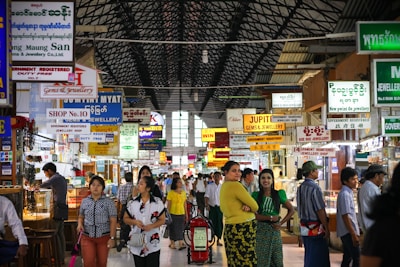 A bustling indoor market with numerous jewelry shops. Various signs written in different languages, including English, hang from the ceiling advertising various shops. The market is crowded with people, some standing and others walking. The architecture features a high, arched ceiling with a lattice design.