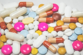 A variety of colorful pills and capsules are scattered across a surface. The assortment includes round, oval, and cylindrical shapes in shades of white, pink, yellow, orange, red, and blue.