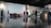 A spacious indoor area with a polished floor reflecting the scene. Several people are scattered throughout, some seated on a red couch and others walking or standing. The backdrop features a large wall labeled 'Gallery' and 'NFT3 delegate centre'. Stairs lead to an upper level, and the setting appears to be a modern public or cultural space.