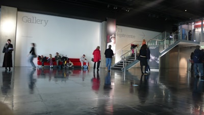 A spacious indoor area with a polished floor reflecting the scene. Several people are scattered throughout, some seated on a red couch and others walking or standing. The backdrop features a large wall labeled 'Gallery' and 'NFT3 delegate centre'. Stairs lead to an upper level, and the setting appears to be a modern public or cultural space.