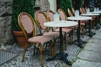 A row of outdoor cafe tables with white tabletops and metal bases lining a cobblestone sidewalk. Each table is paired with woven rattan chairs featuring a red and white checkered pattern. Tall potted green plants are placed between some tables, and the setting is bordered by a stone wall.