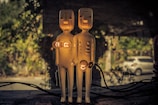 Two figurine lamps with humanoid shapes stand side by side on a surface. They have switches for faces, and light bulbs are mounted at their chests and in the pelvic region. The background contains blurred greenery and a parked car, suggesting an outdoor setting with wires visible near the bottom of the image.