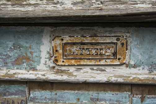 A weathered and rusty metal letters slot is mounted on a door with peeling paint and a distressed wood surface. The surrounding wood exhibits patches of chipped turquoise paint and exposed raw wood, giving it a rustic and aged appearance.