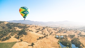 A vibrant hot air balloon floats over a sprawling, hilly landscape. The terrain features golden hills dotted with patches of green trees and small bodies of water. In the distance, blue mountains rise under a clear blue sky.